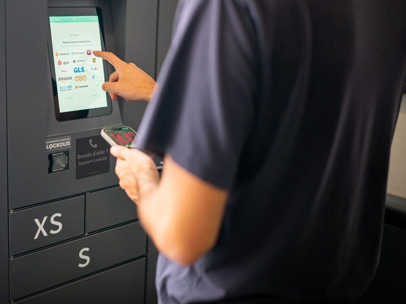 Un livreur, smartphone en main, choisit son transporteur sur l'écran tactile d'un casier à colis Lockolis